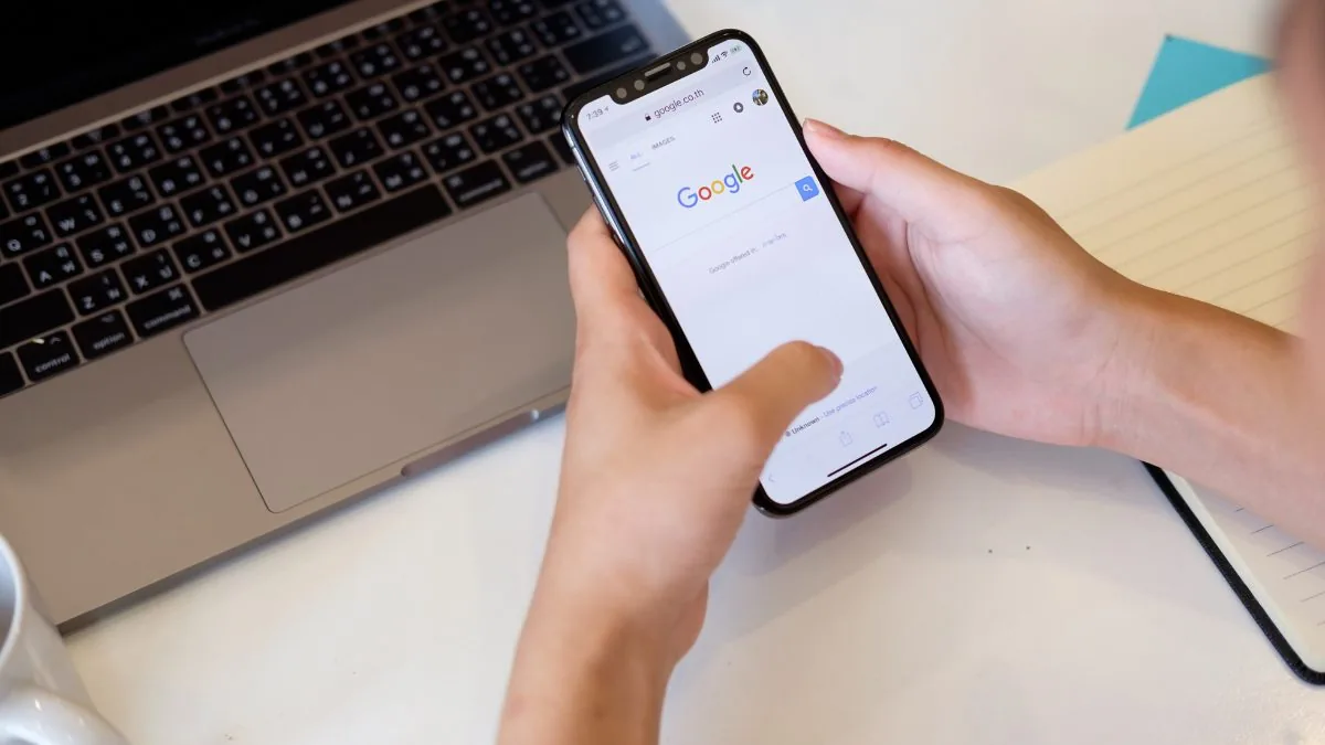 Mãos segurando um celular com a busca do Google aberta, ao lado de um notebook